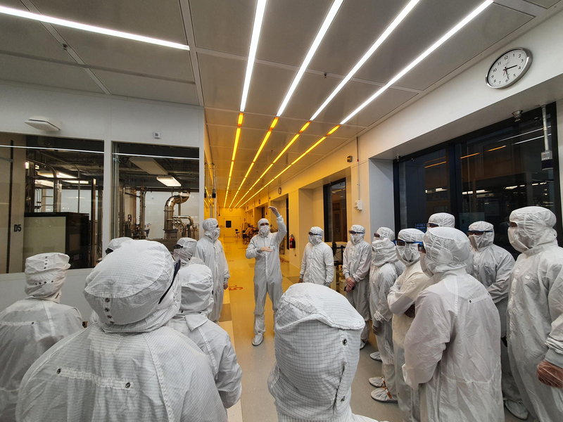 A view from behind of about 15 people dressed head-to-toe in white cleanroom suits, facing another gowned-up individual gesturing as they speak.