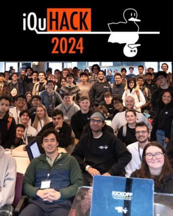 Perhaps 150 people pose for a photo in a large classroom. About a third are seated in the foreground and the rest stand. At top is a banner that reads iQuHACK 2024, with the image of two nearly identical ducks, one alive and one shown as dead with X's for eyes