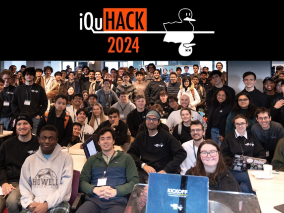 Perhaps 150 people pose for a photo in a large classroom. About a third are seated in the foreground and the rest stand. At top is a banner that reads iQuHACK 2024, with the image of two nearly identical ducks, one alive and one shown as dead with X's for eyes