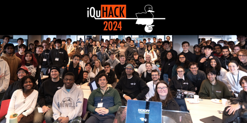 Perhaps 150 people pose for a photo in a large classroom. About a third are seated in the foreground and the rest stand. At top is a banner that reads iQuHACK 2024, with the image of two nearly identical ducks, one alive and one shown as dead with X's for eyes