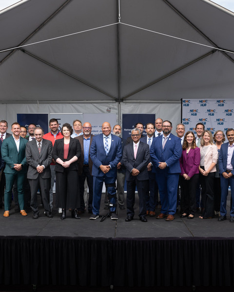 36 individuals pose on an outdoor stage under a pyramidal awning
