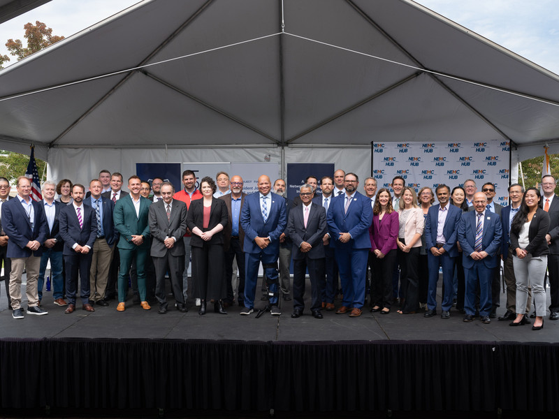 36 individuals pose on an outdoor stage under a pyramidal awning