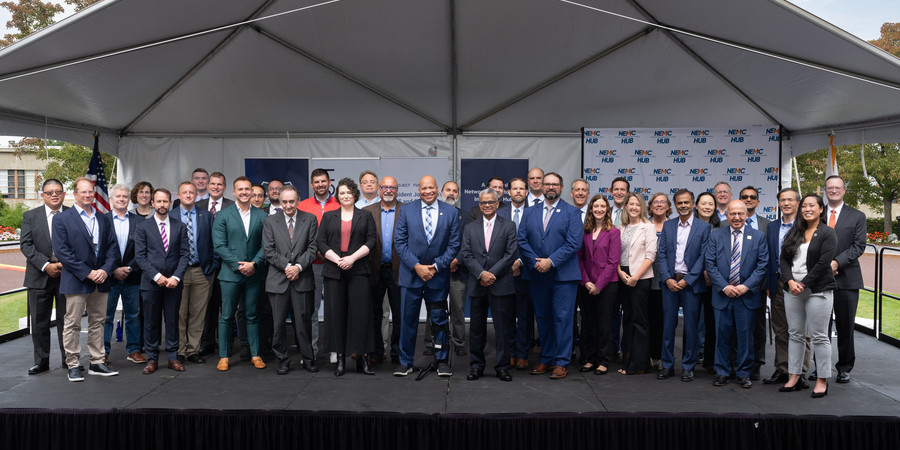 36 individuals pose on an outdoor stage under a pyramidal awning