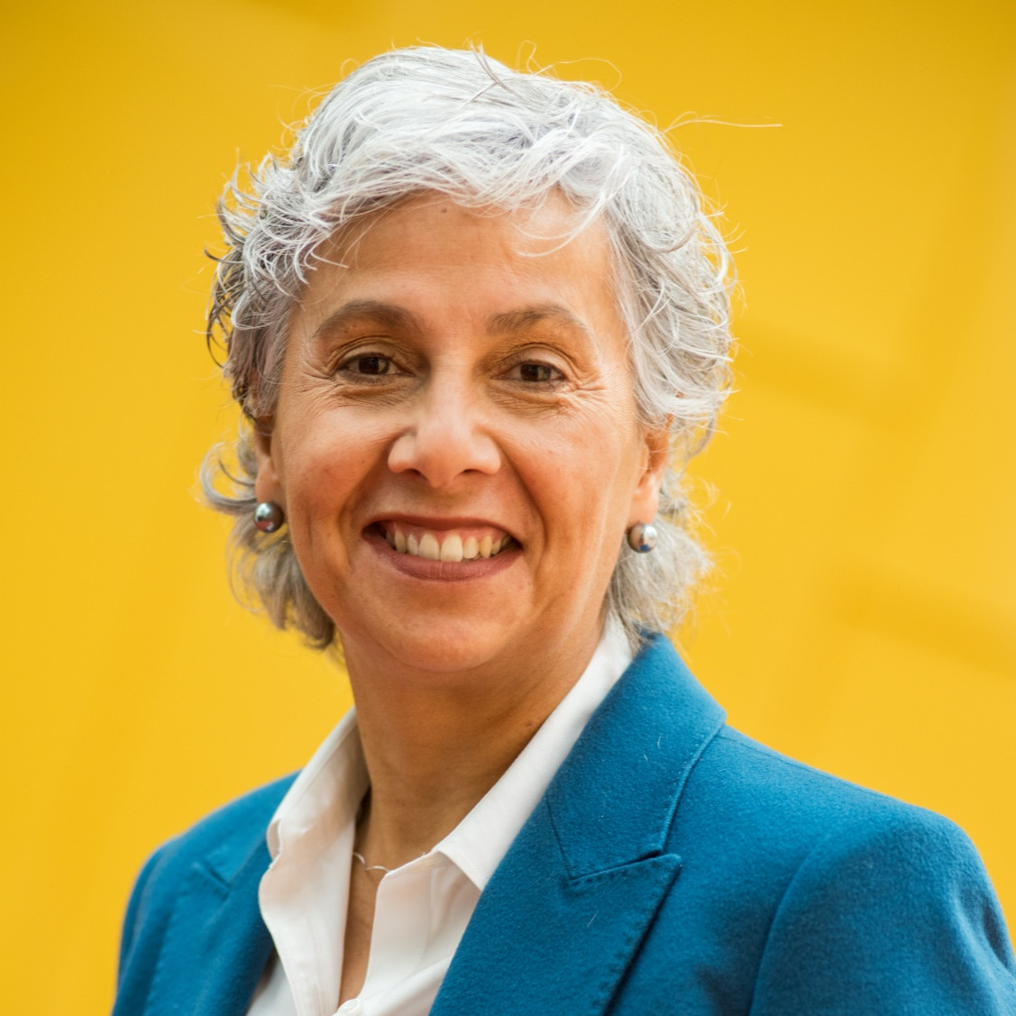 headshot of Una-May wearing white shirt and bright blue suit jacket on a bright yellow backdrop
