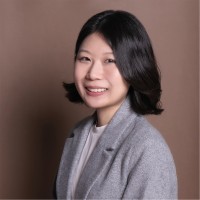woman with short dark hair wearing a white shirt and great coat in front of a light brown background