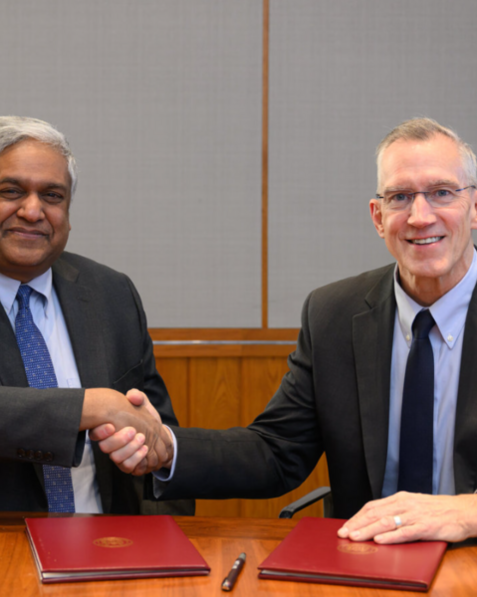 Anantha Chandrakasan and Gregg Bartlett, both in suits, sit at a table and shake hands for the camera
