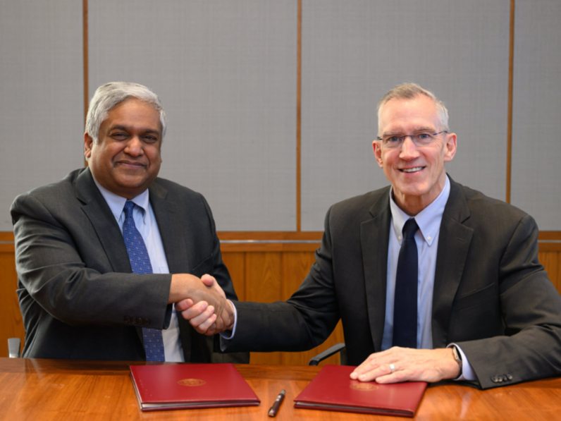 Anantha Chandrakasan and Gregg Bartlett, both in suits, sit at a table and shake hands for the camera