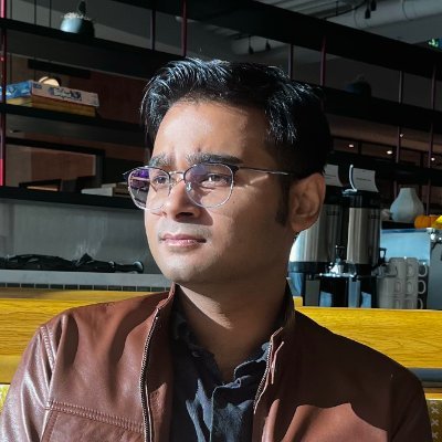 headshot of male wearing wire rim glasses and brown leather jacket seated on wood bench