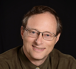 male with glasses wearing brown dress shirt in front of black background