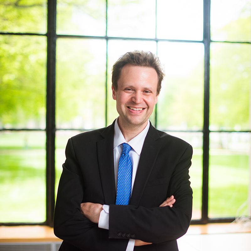 man wearing black suit, white shirt, and blue tie standing in front of window