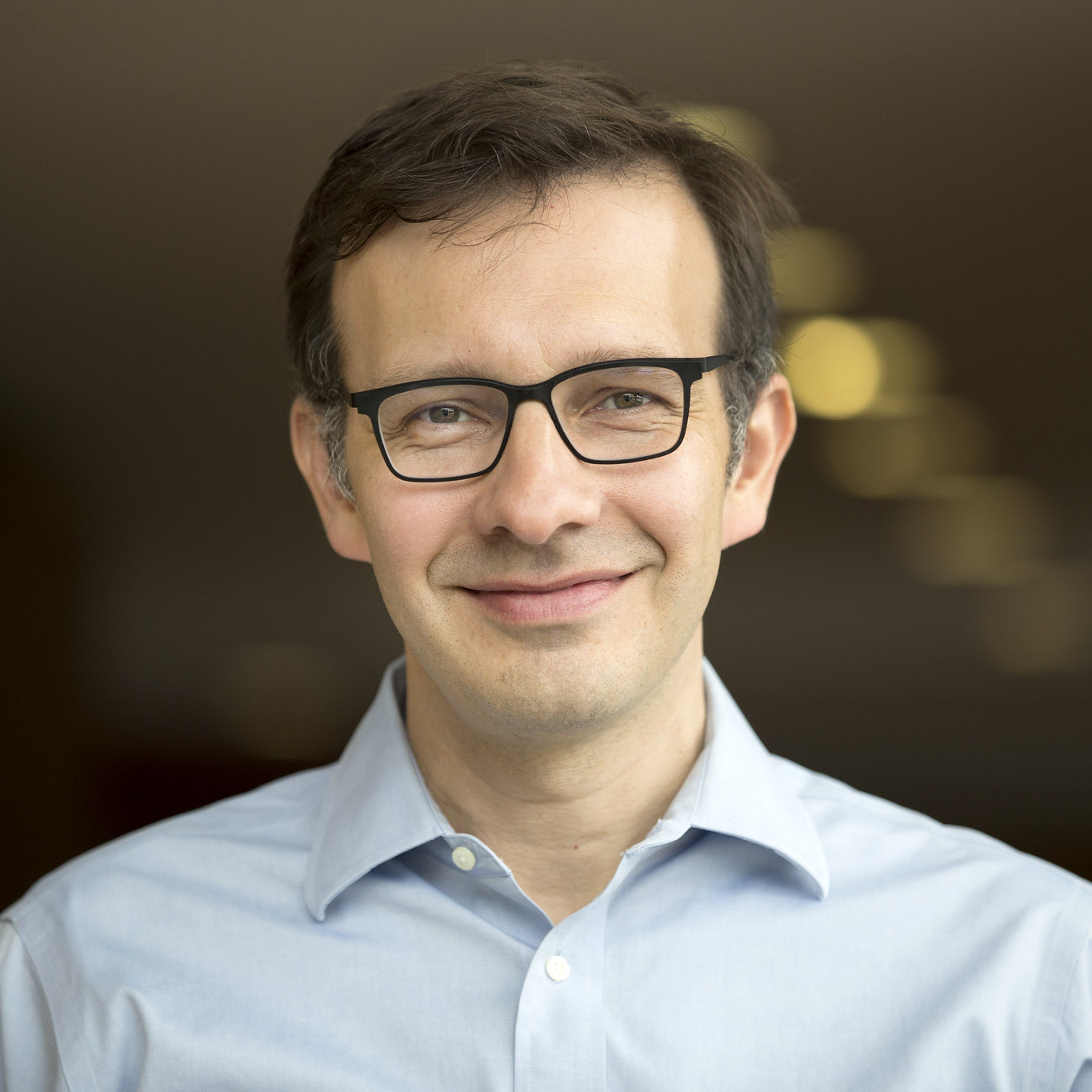 man with brown hair wearing glasses and blue collared shirt in front of blurred brown background