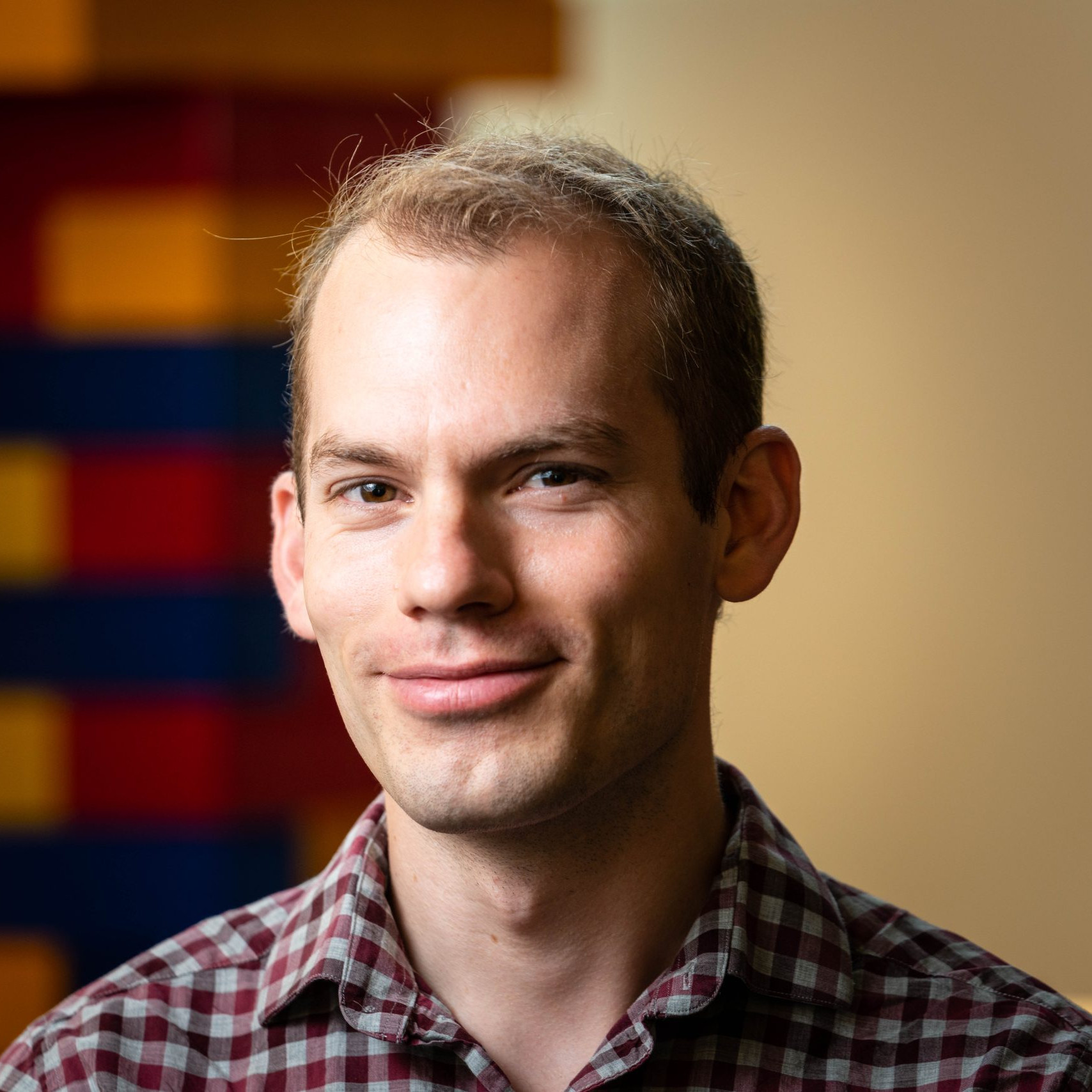 man wearing checkered shirt in front of blurred tan, yellow, blue, and red background