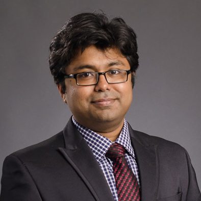man with dark hari and glasses wearing a suit in front of a grey background
