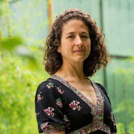 woman with brown hair in a black dress standing in front of a glass building and green trees