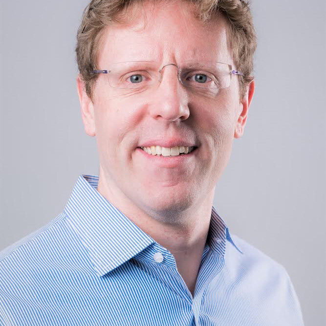 man with brown hair, glasses, and wearing blue collared shirt in front of grey background