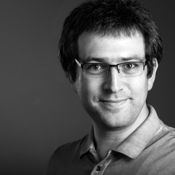 man with dark hair and glasses wearing collared shirt in front of grey background