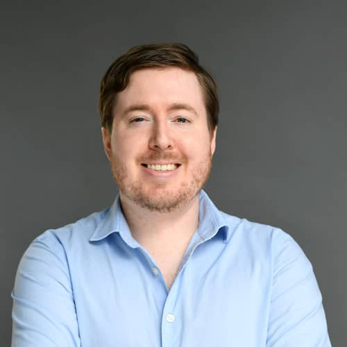male with brown hair wearing light blue dress shirt in front of grey backdrop